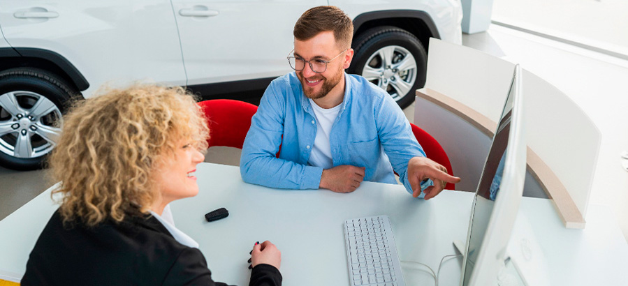 Um homem e uma mulher conversando em um ambiente de vendas de automóveis, com um computador e um carro ao fundo, transmitindo um clima de atendimento amigável e profissional para consórcio ou financiamento.