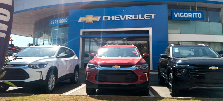 Imagem de uma concessionária Chevrolet com três veículos em exibição: SUV branca, vermelha e preta, destacando a marca e o logo Chevrolet.